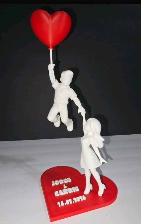 Figura impresa en 3D de una pareja con un globo en forma de corazón, sobre una base roja con sus nombres y la fecha